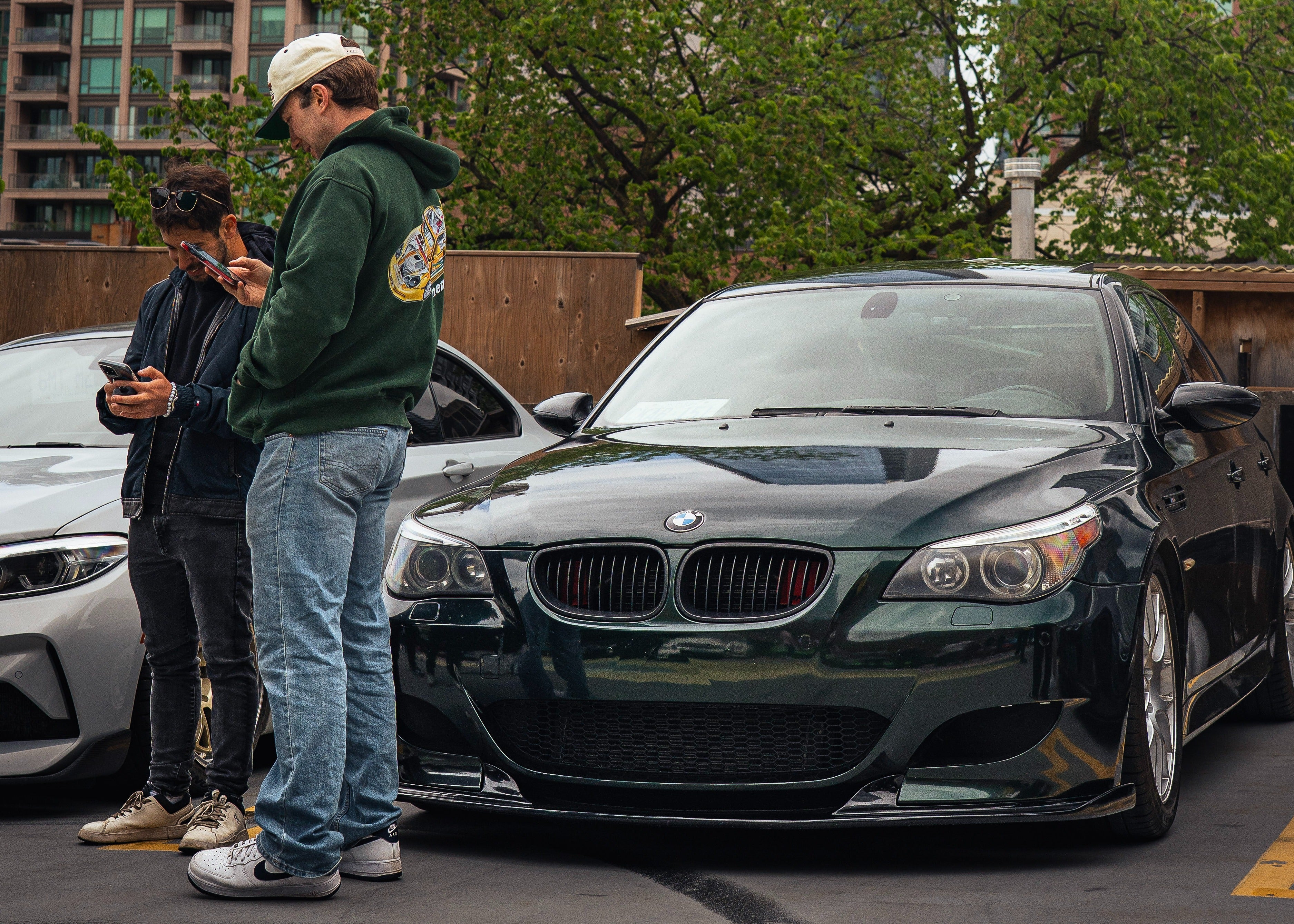 Two people standing next to a black BMW car in an urban setting with tall buildings and trees in the background.
