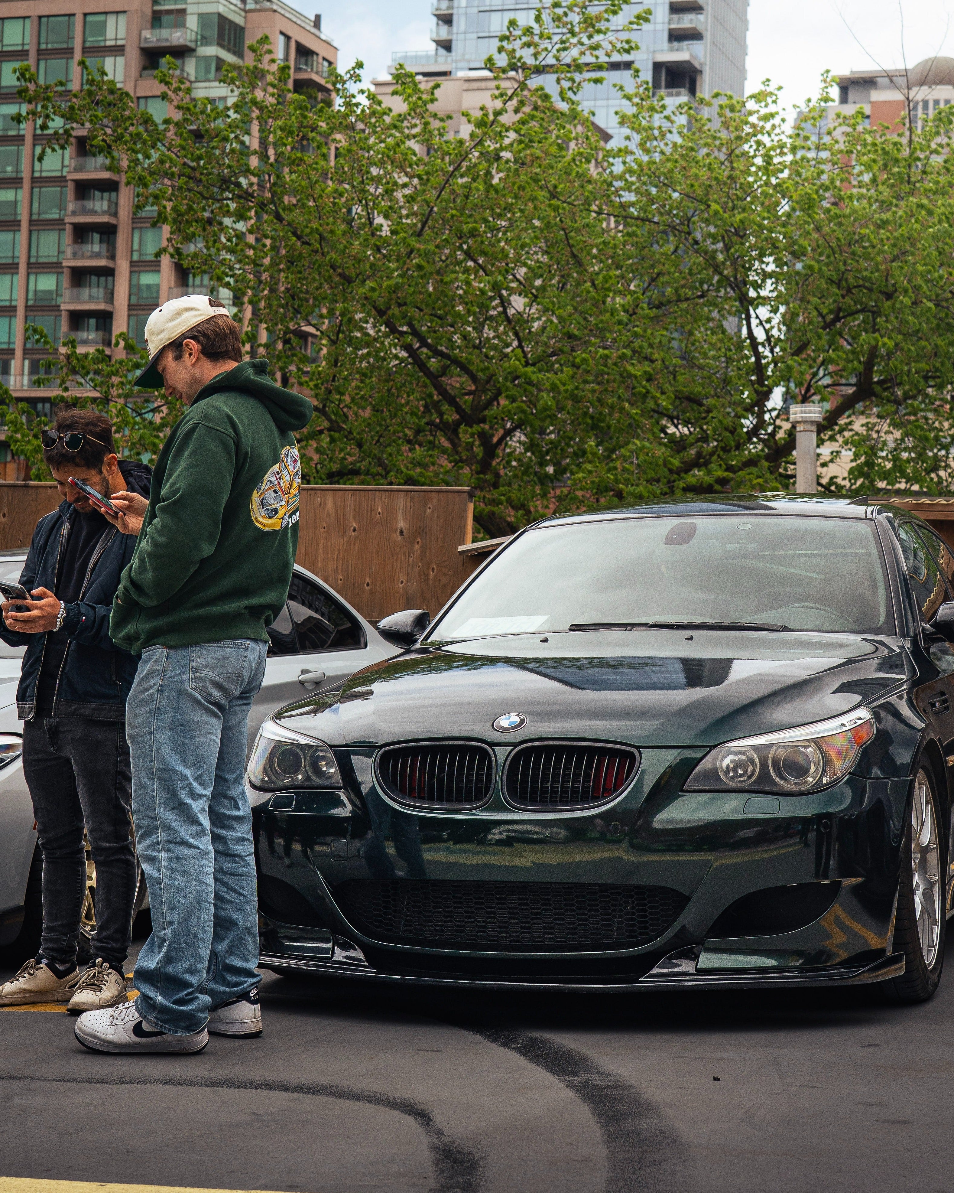 Two people standing next to a black BMW car in an urban setting with tall buildings and trees in the background.